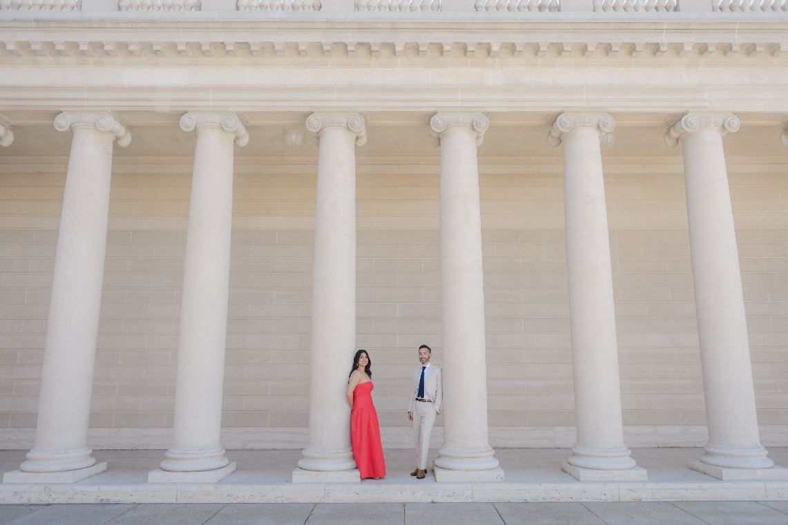 Pooja in a pink dress and Derrick standing among the columns of the Legion of Honor