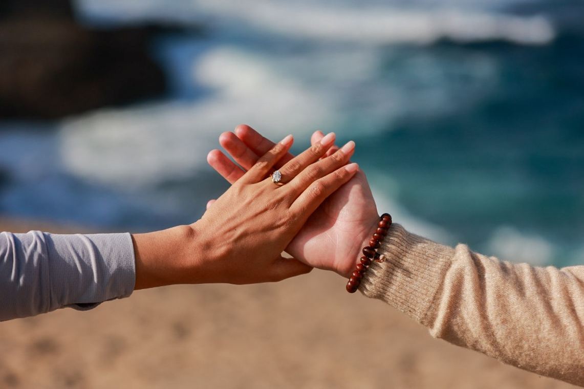 Their hands clasped above the sand, showing the engagement ring