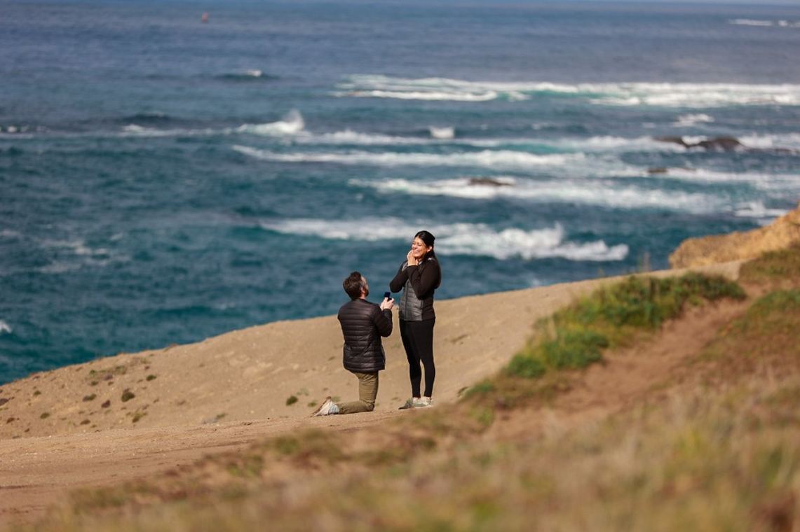 Derrick on one knee proposing to Pooja on a coastal bluff